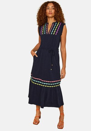 Moteris su garbanotais plaukais, dėvinti be rankovių tamsiai mėlyną suknelę su spalvotomis banguotomis juostomis ir oranžinius basutes su dirželiais, stovinti viena ranka kišenėje.