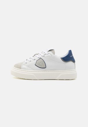 Zapatillas de cuero blancas con puntera de ante gris, acento en el talón en azul marino, parche de logo texturizado en el lateral y suela gruesa de goma blanca.