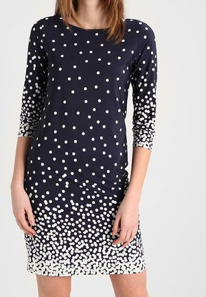 Femme vêtue d'une robe bleu marine à manches trois quarts avec des pois blancs dont la densité augmente vers l'ourlet.