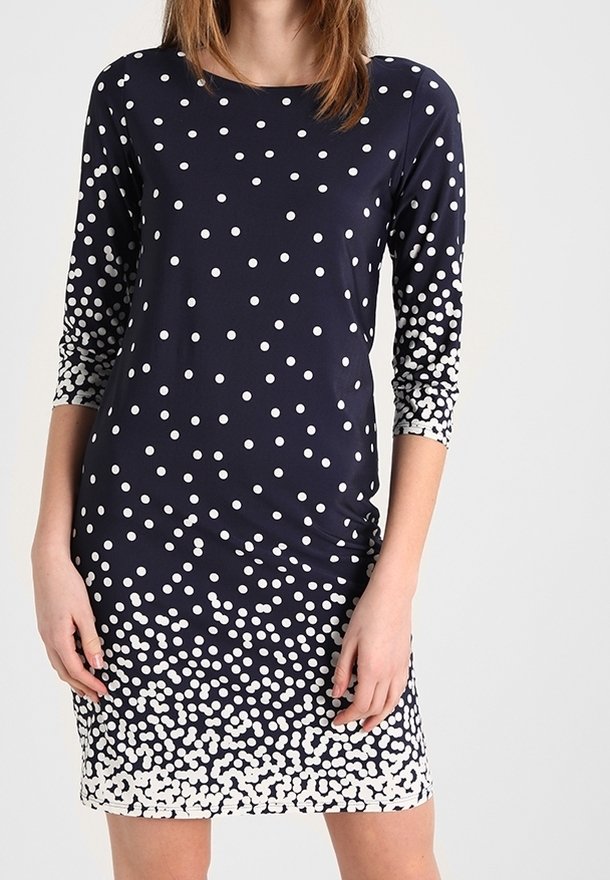 Femme vêtue d'une robe bleu marine à manches trois quarts avec des pois blancs dont la densité augmente vers l'ourlet.