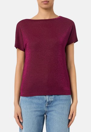 Donna che indossa una maglietta a maniche corte scintillante di colore magenta scuro abbinata a jeans denim azzurri chiari, in piedi davanti a uno sfondo semplice.