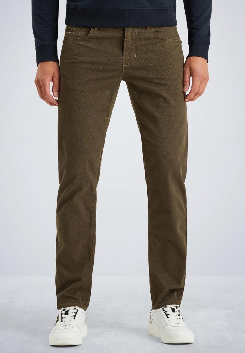 Homme portant un pantalon droit vert olive et des baskets blanches, debout devant un fond clair et uni, les mains détendues sur les côtés.