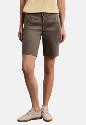 Modelka w brązowych szortach do kolan, jasnobeżowych casualowych sneakerach i wpuszczonej w spodnie bladożółtej koszuli, stojąca na białym tle.