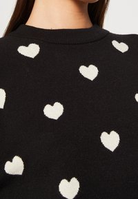 Schwarzer Pullover mit Rundhalsausschnitt, der gewebte weiße Herzmuster auf dem Stoff aufweist, eine weiche Textur hat und eine taillierte Silhouette besitzt.