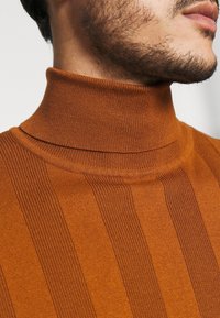 Gros plan sur le bas du visage et le cou d’un homme portant un pull à col roulé côtelé orange brûlé.