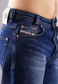 Nahaufnahme einer Person, die blaue Jeans trägt, die die Vorderseite und die kleine Münztasche mit Etikett und Nähdetails zeigt.