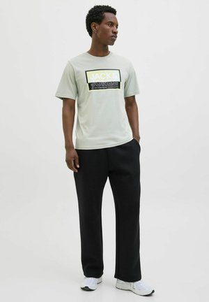 Lichtgroene T-shirt met korte mouwen met een gedurfde zwart-gelbe graphic print. Zwarte relaxte broek en witte sportschoenen maken de outfit compleet.