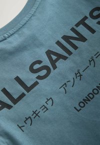 Sininen puuvillainen t-paita, jossa rohkea musta teksti. Kaula-aukko on pyöreä, ja kangas on sileä ja hienovaraisesti kiiltävä.