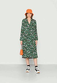 Vestido floral verde com mangas longas e decote em V, combinado com um chapéu de balde laranja, sapatilhas pretas e uma pequena bolsa laranja.