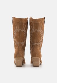 Botas de vaquero de ante color canela con tacón medio, que presentan patrones de estallido de estrellas bordados en blanco en la parte trasera y lengüetas para facilitar su uso.