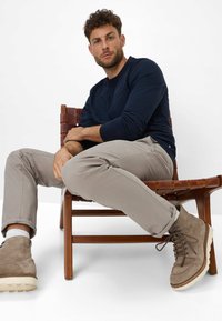 Navy long-sleeve shirt and light beige trousers sit on a wooden chair. Brown suede boots feature lacing and white soles.