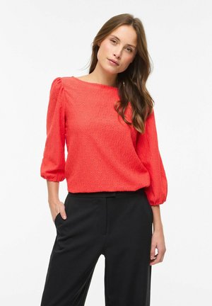 Ženska z dolgimi rjavimi lasmi, oblečena v teksturirano koralno bluzo z nabranimi rokavi in črne hlače, stoji z eno roko v žepu.