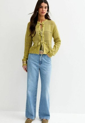 Femme portant un jean taille haute bleu clair à jambes larges et un cardigan vert en crochet noué avec des nœuds, debout devant un fond blanc.
