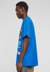 Blå bomulds t-shirt med en afslappet pasform, prydet med grafiske prints på forsiden i forskellige farver. Korte ærmer og en rund udskæring er synlige.