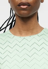 Κοντινό πλάνο ενός ατόμου με πλεξούδες που φοράει ένα τοπ σε μέντα πράσινο χρώμα με ζιγκ-ζαγκ σχέδιο και στρογγυλή λαιμόκοψη.