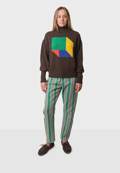 Maglione di colore marrone lavorato a maglia con un design geometrico in verde, giallo e rosso. Abbinato a pantaloni a righe verdi e scarpe di colore marrone scuro.
