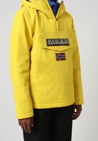 Gul vandtæt jakke med hætte, der har en frontlomme, mærkeetiket og et lille patch med det norske flag på venstre side.