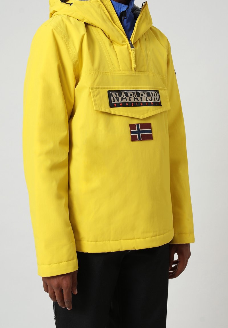 Gul vandtæt jakke med hætte, der har en frontlomme, mærkeetiket og et lille patch med det norske flag på venstre side.