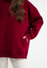 Bordeaux hoodie met een voorzak, losse pasvorm, geribde manchetten en zoom. De stof lijkt zacht met een gladde textuur tegen een witte achtergrond.