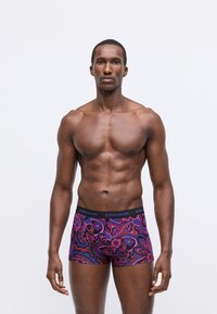 Svalnatý muž s tmavou pokožkou stojí čelom dopredu, oblečený v farebných boxerách s vzorom a čiernym pásom so značkou "BJÖRN BORG."