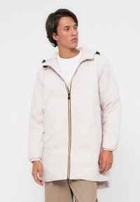Manteau long rose clair avec une capuche en fourrure, fermeture éclair sur le devant, poignets élastiques et un accent de fermeture éclair multicolore. Texture de tissu lisse.