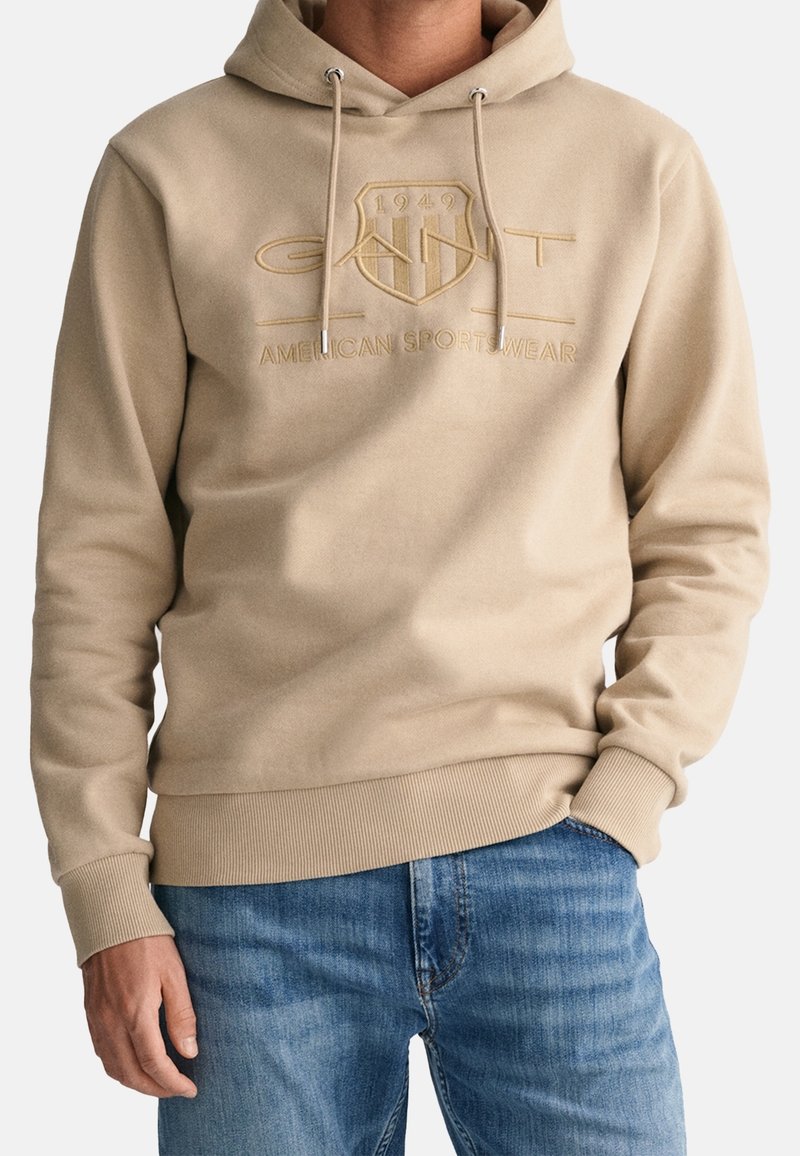 Sudadera beige con cordones, que presenta un logo en relieve con la inscripción "GANT 1949 AMERICAN SPORTSWEAR" y puños acanalados. Llevada con pantalones vaqueros azules.