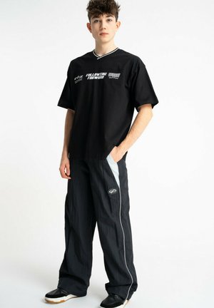 Młody mężczyzna w czarnej koszulce z nadrukiem i luźnych czarnych spodniach z białym paskiem, stojący z jedną ręką w kieszeni na białym tle.