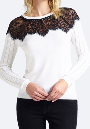Pull blanc avec une superposition de dentelle noire aux épaules, présentant un bord en forme de vagues et des manches à côtes verticales. Les accents comprennent de petites clous.
