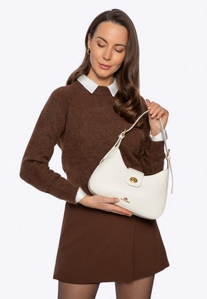 Vrouw in een bruine trui en rok met een witte leren handtas met gouden gesp en logo tegen een effen achtergrond.