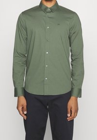 Man in een slim-fit olijfgroene overhemd met lange mouwen en zwarte broek, staande tegen een effen achtergrond.