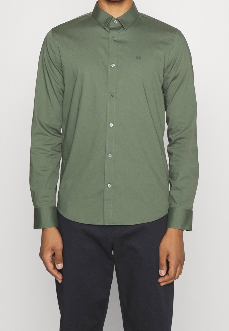 Man in een slim-fit olijfgroene overhemd met lange mouwen en zwarte broek, staande tegen een effen achtergrond.