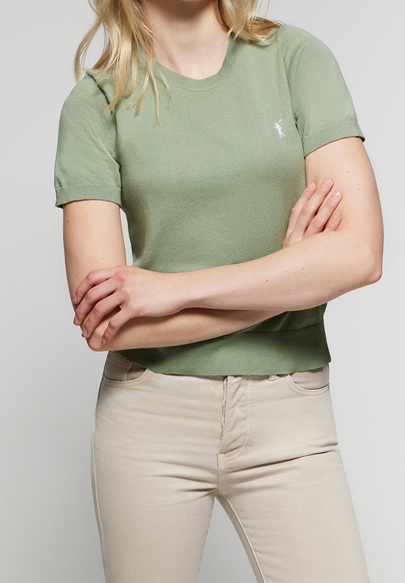 Haut tricot vert à manches courtes avec un col rond, agrémenté d'un petit logo blanc sur la poitrine. Assorti à un pantalon beige.