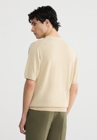 Maglione corto a maniche corte beige in maglia con scollo e orlo a coste, caratterizzato da una texture liscia e una silhouette aderente.