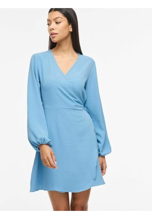 Žena v dlhých rukávoch, svetlomodré zavinovacie šaty stojaca pred jednoduchým bielym pozadím.