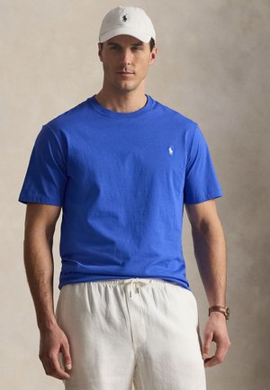 Mand iført en blå kortærmet skjorte, hvide bukser med snøre, hvid kasket og brun armbåndsur foran en neutral baggrund.