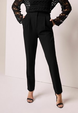 Black tailored pants with a high waist, pleats, and a straight leg design. Worn with a textured black lace top and open-toe black heels.