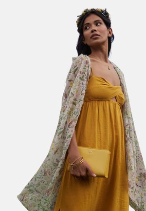 Vestido amarillo maxi con un escote anudado y cintura fruncida, combinado con un chal transparente de patrón floral, sosteniendo un clutch amarillo.