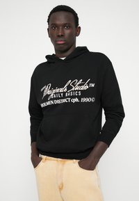 Μαύρο φούτερ με τσέπη μπροστά, με λευκό κεντημένο κείμενο. Φορεμένο με ανοιχτόχρωμο τζιν. Απλός σχεδιασμός, ελαστικές μανσέτες και κοπή στο κάτω μέρος.