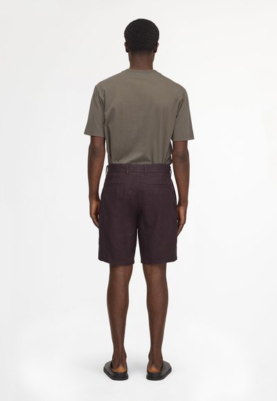 Homme debout de dos portant une chemise à manches courtes vert olive, un short marron foncé jusqu'aux genoux et des sandales noires.