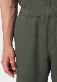 Pantaloni in lino verde oliva con vita elastica, tasche laterali e una consistenza liscia con dettagli di cucitura sottili.