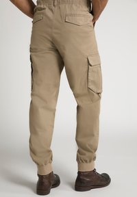 Pantaloni cargo beige con elastico in vita, tasche laterali e posteriori, polsini affusolati e una texture liscia, abbinati a scarpe marroni scure.