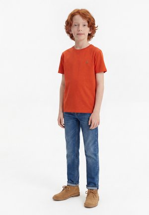 Rudy chłopiec stoi, ubrany w pomarańczowy t-shirt z krótkim rękawem, niebieskie dżinsy oraz jasnobrązowe sznurowane buty na białym tle.