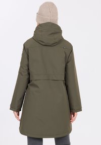 Olivgrön vattentät lång jacka med huva, som har bältedetalj och en avslappnad passform. Stickad beige mössa bärs på huvudet.