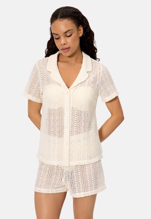 Mujer vistiendo un top blanco de manga corta con botones y tejido abierto, y pantalones cortos de punto a juego, sobre un fondo liso.