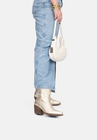 Gold metallic western-style ankle boots with pointed toes and stacked heels, paired with light blue denim, holding a small white handbag.