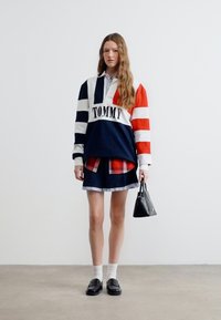 Γυναίκα που φοράει φούτερ Tommy με ναυτικό, άσπρο και κόκκινο ριγέ, καρό φούστα, άσπρες κάλτσες, μαύρα μοκασίνια και κρατάει μια μικρή μαύρη τσάντα χειρός.