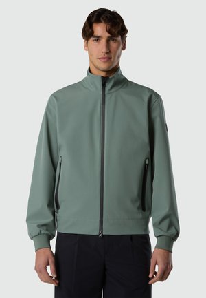 Man wearing a sage green zip-up jacket with black zipper details and black pants, standing against a plain gray background.