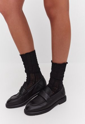 Ben klædt i strukturerede sorte ankelstrømper parret med sorte læder loafers mod en simpel lys baggrund.