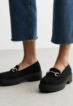 Black platform loafers with a textured suede finish, featuring a gold metal accent and thick black sole. Pairing with denim cropped pants.