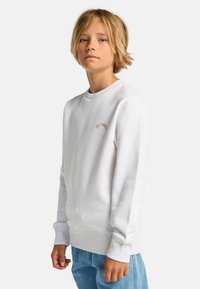 Felpa bianca in cotone con polsini e orlo a costine, caratterizzata da un piccolo logo beige sul petto. Abbinata a jeans in denim blu chiaro.